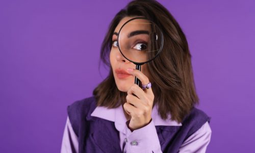 Young woman in shirt and vest on purple background having fun with magnifying glass in hand
